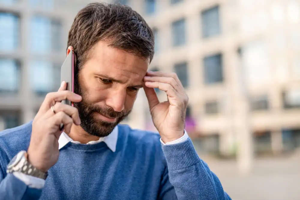 Bearded man in a blue sweater on a cellphone outdoors, hand to temple, looking worried or focused.