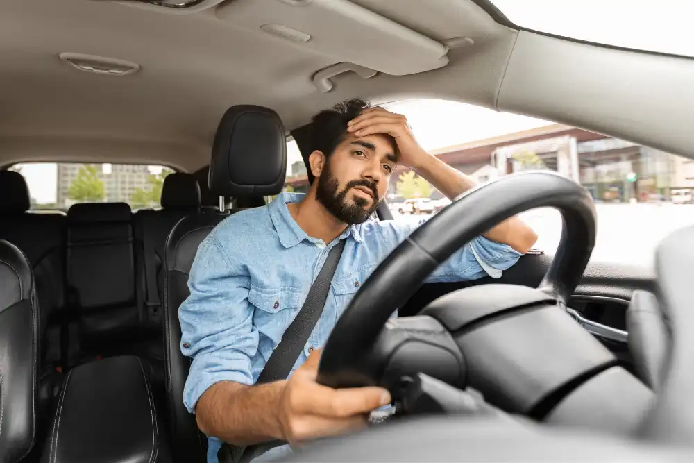 Bearded man in a denim shirt grips the steering wheel with one hand on his head, looking stressed while driving in city traffic to the right of the frame.