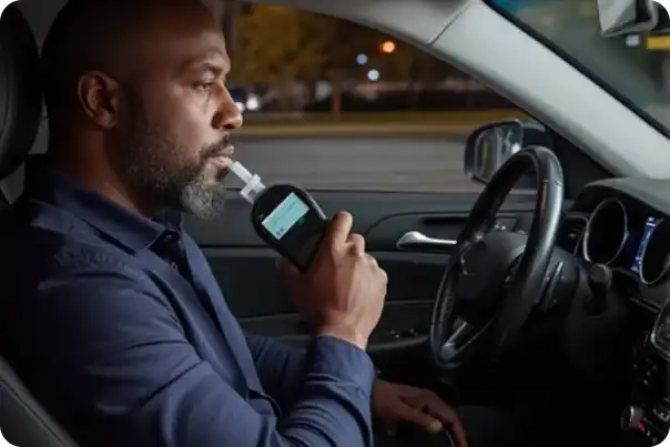 Man sitting in a car, blowing into a breathalyzer device mounted to a handheld reader on the center console (in-car sobriety test).