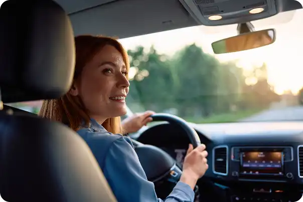 Woman driving a car, smiling while holding the steering wheel in the driver’s seat at sunset light.