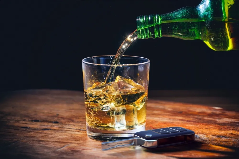Whiskey being poured into a glass with ice on a wooden table, with a car key fob resting nearby.