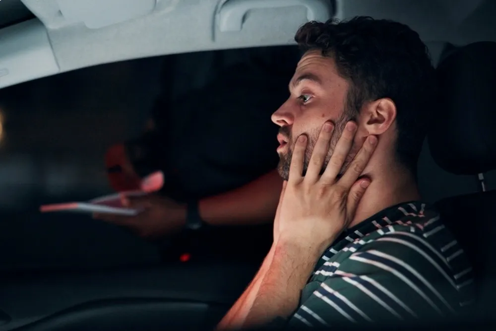 Man in a car, gripping his throat with both hands and looking startled, with a second person in the background holding a card flank.