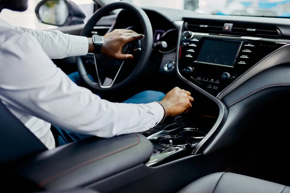 Person in a white shirt driving a car, hands on the steering wheel inside a modern dashboard.
