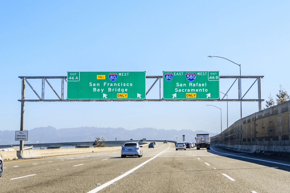 Green overhead highway signs on a multi-lane freeway directing to San Francisco Bay Bridge (I-80 West) and San Rafael/Sacramento (I-80 East, I-580 West) with multiple vehicles under a blue sky.