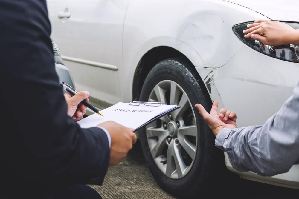 Person in a suit writes on a clipboard while inspecting a dented white car beside the tire.