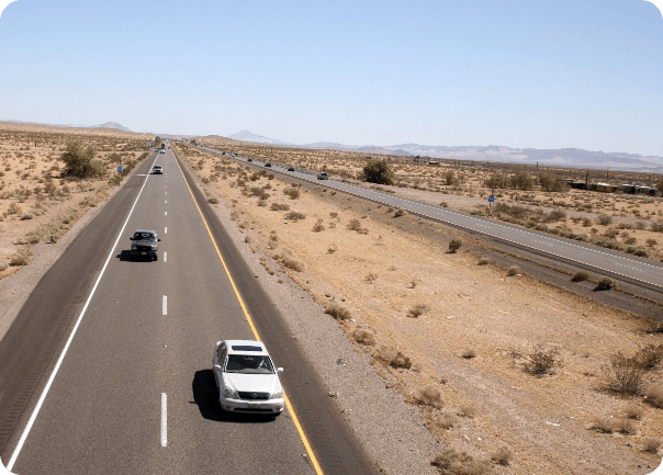 Two-lane desert highway with a white car in the foreground and other vehicles in the distance, scrubby terrain and distant mountains under a clear blue sky.