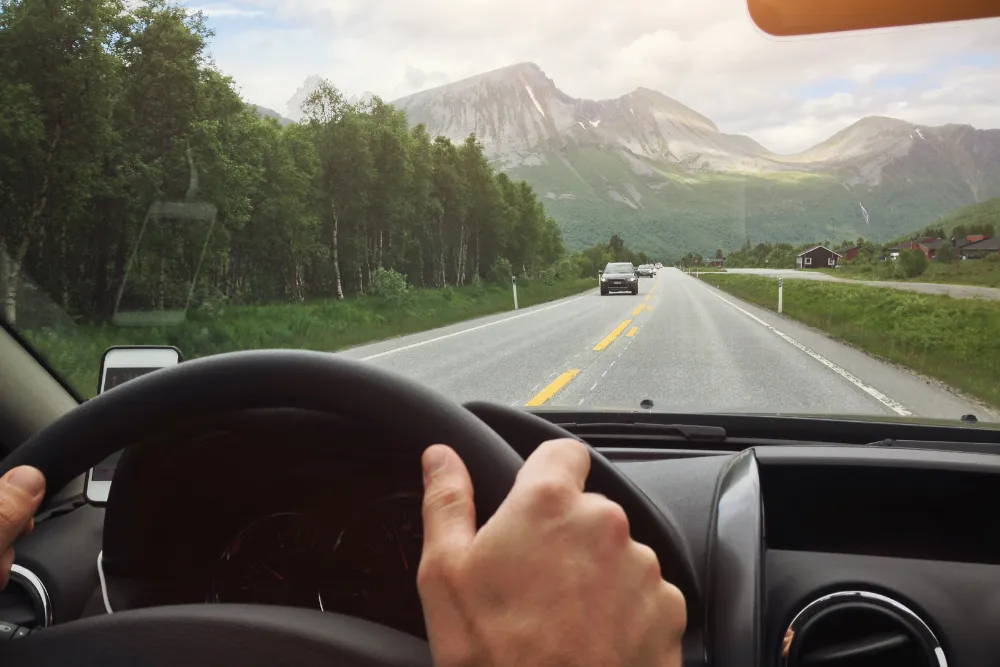 View from inside a car: hands on the steering wheel driving toward a two-lane road with mountains in the distance.